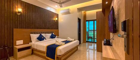 A modern hotel room with a wooden headboard, a flat-screen TV, a ceiling fan, and a view of the outdoors through a large window.