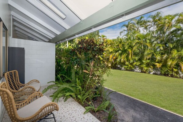 Un patio al aire libre con mobiliario de mimbre, piso de baldosas y vistas a un césped cuidado y plantas tropicales.