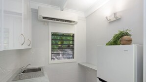 Cocina compacta con armarios blancos, pileta integrada, ventana con persianas, aire acondicionado y refrigerador blanco.