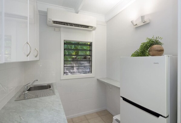 Cocina compacta con armarios blancos, pileta integrada, ventana con persianas, aire acondicionado y refrigerador blanco.