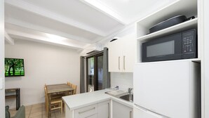 Een compacte keuken met witte kastjes, een witte koelkast, een magnetron en een eethoek met een houten tafel en stoelen.