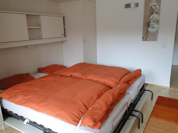 Una cama individual con una manta y almohadas anaranjadas, una mesita de noche pequeña, un estante en la pared y un cuadro de flores.