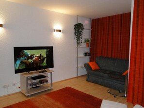 A living room with a TV, a grey sofa, a red curtain, and a plant on a shelf.