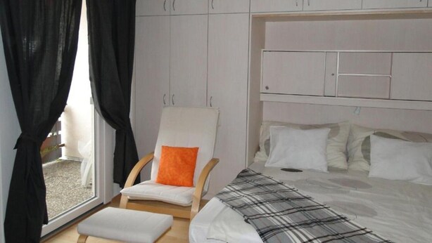 A bedroom with a bed, a chair with an orange cushion, a window with black curtains, and a white wardrobe. - Apartment 'Studio in Sankt Blasien' with Balcony and Wi-Fi (St Blasien)
