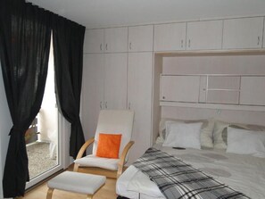 A bedroom with a bed, a chair with an orange cushion, a window with black curtains, and a white wardrobe.