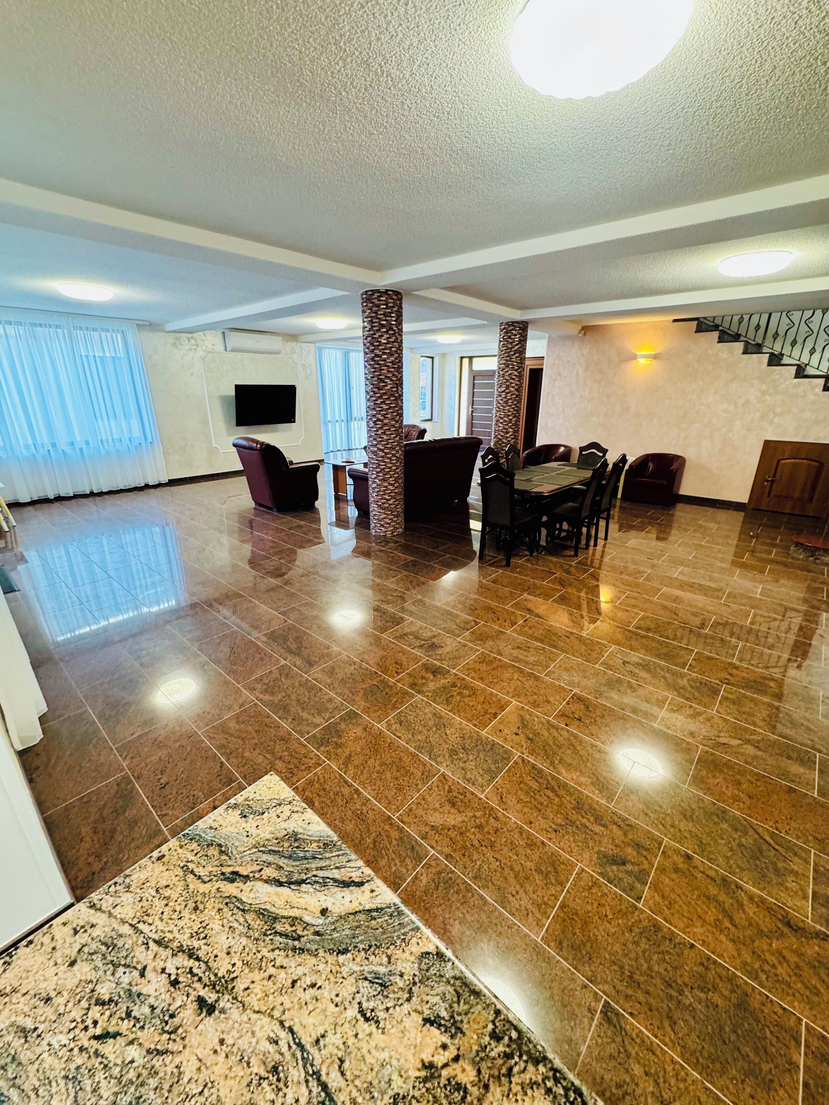 A spacious living area with a polished tile floor, a dining table with chairs, a sofa, and a staircase with a railing.