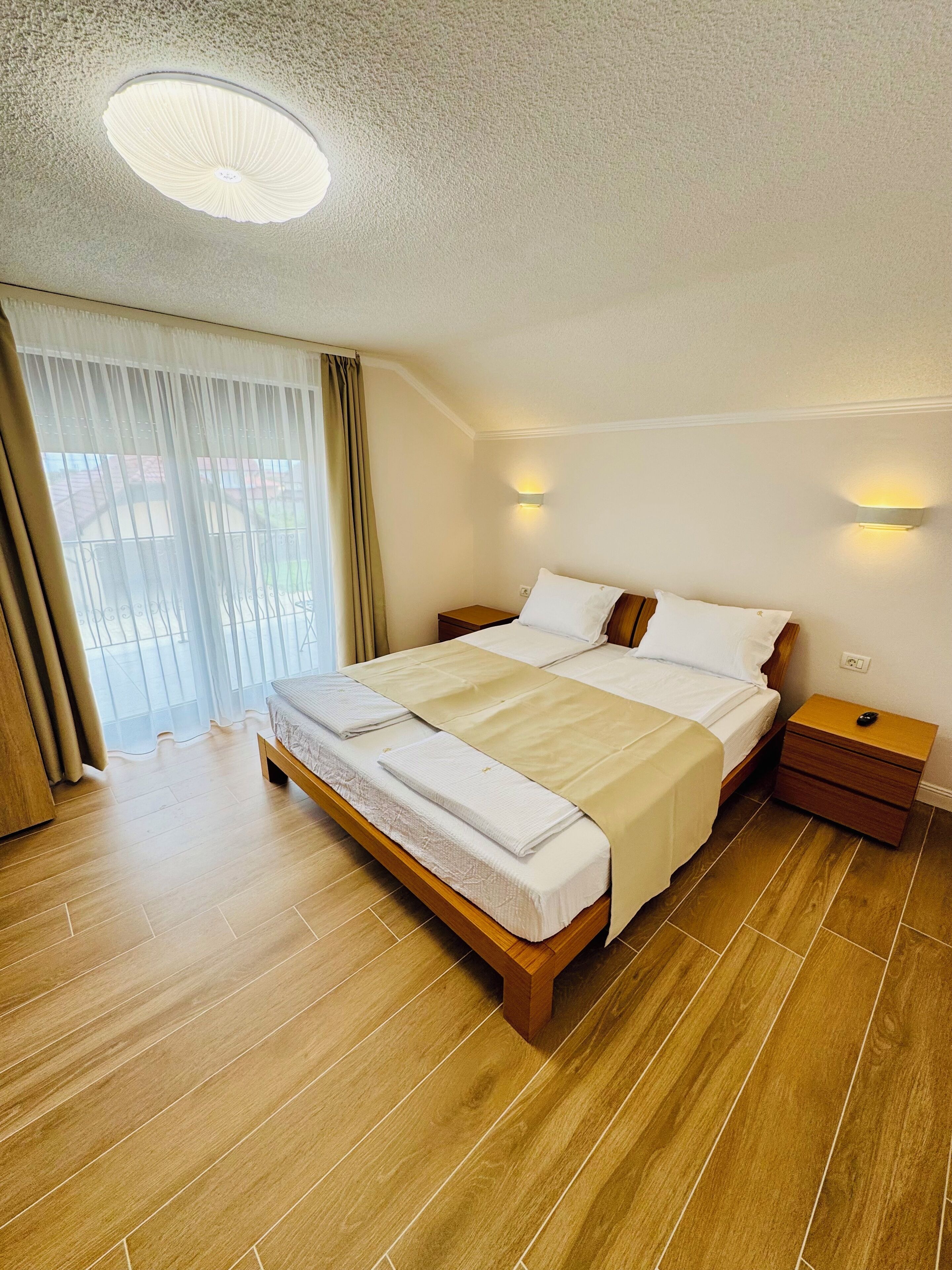 Habitación de hotel con una cama grande, suelo de madera, una lámpara de techo y una ventana con cortinas.