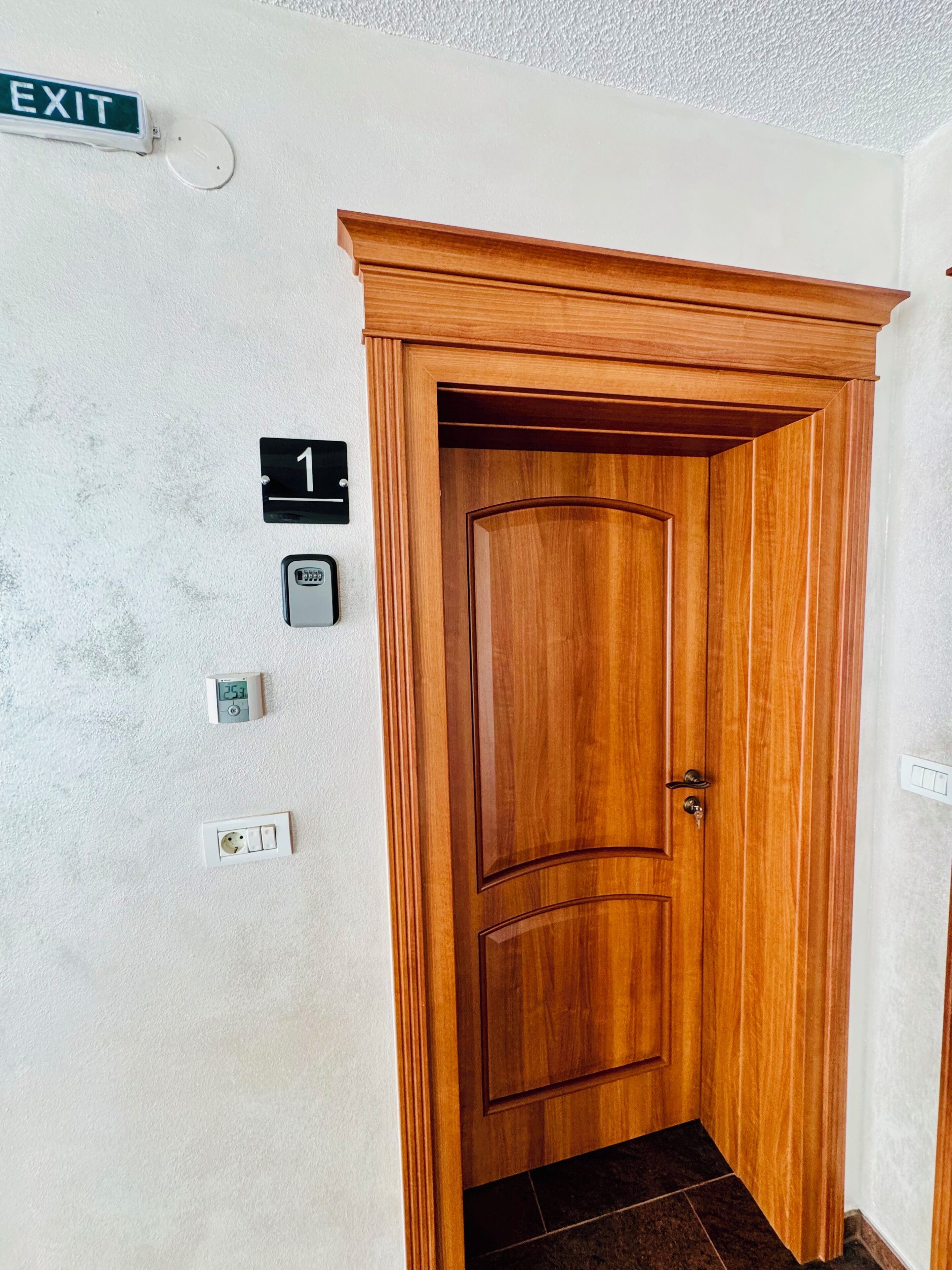 Une porte en bois avec un panneau portant le numéro 1 au-dessus, un panneau indiquant la sortie, un thermostat et un interrupteur d’éclairage sur le mur.