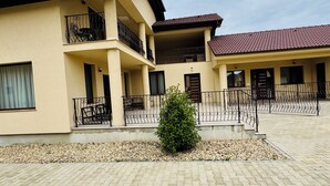 A two-story building with a tiled roof, a balcony with a metal railing, and a paved courtyard with a small bush.