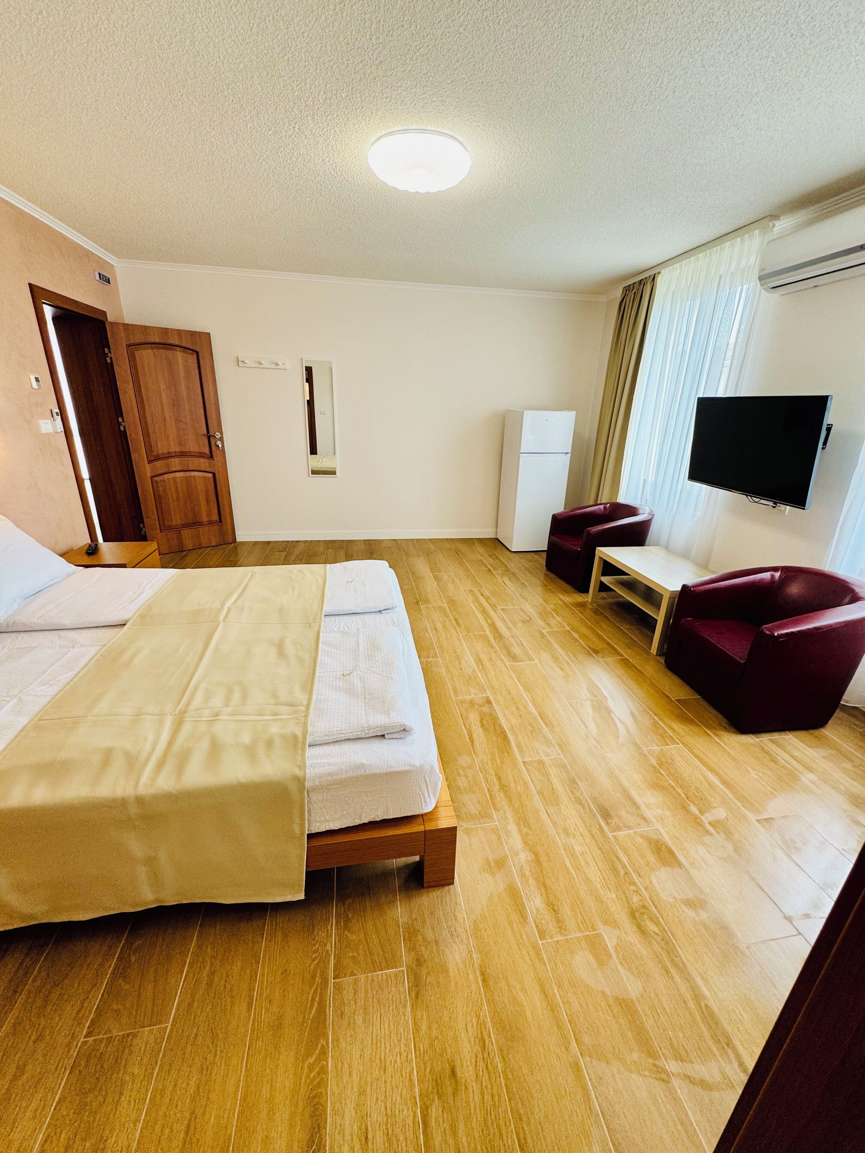 A hotel room with a wooden floor, a bed with a beige bedspread, a wooden wardrobe, a white wall-mounted TV, and a small table with a chair.