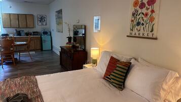 A bedroom with a bed, a bedside lamp, a pillow, a chair, a dresser, and a kitchen area in the background.