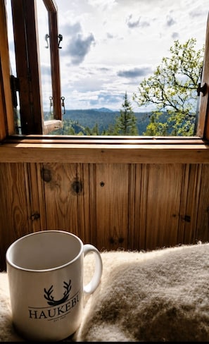 A window with a view of a forest and mountains, a wooden interior, and a white mug with 'HAUKEBU' text.
