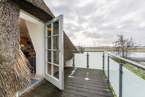 Un bâtiment à toit de chaume, doté d’une porte en verre qui donne sur une terrasse en bois offrant une vue sur une étendue d’eau et sur des éoliennes situées au loin.