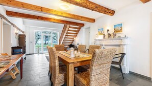 Een eetruimte met een houten tafel en rieten stoelen, een open haard met decoratieve voorwerpen, en een trap die naar een andere kamer leidt.