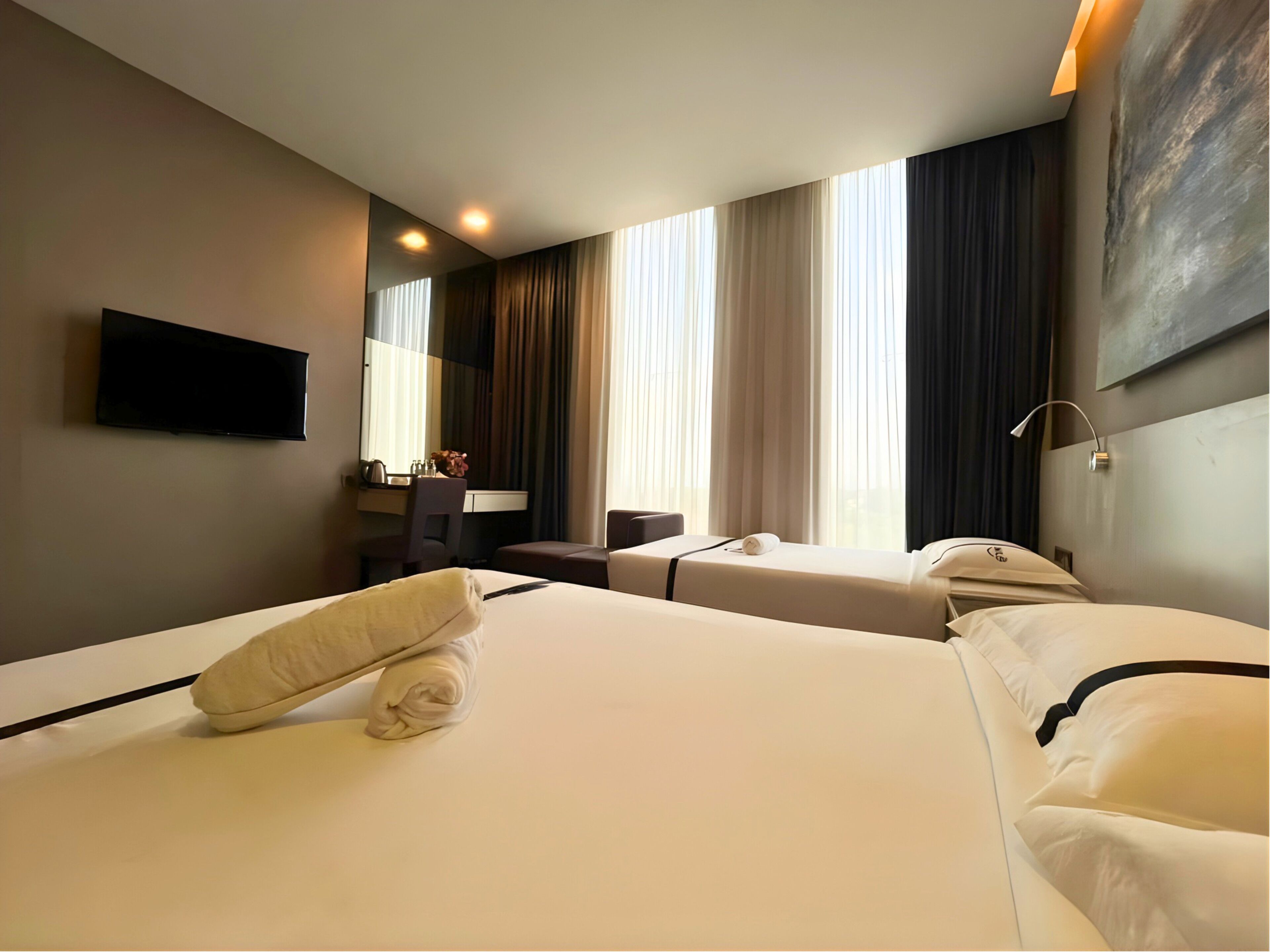 A hotel room with two beds, a desk, a chair, a flat-screen TV, and a large window with curtains.
