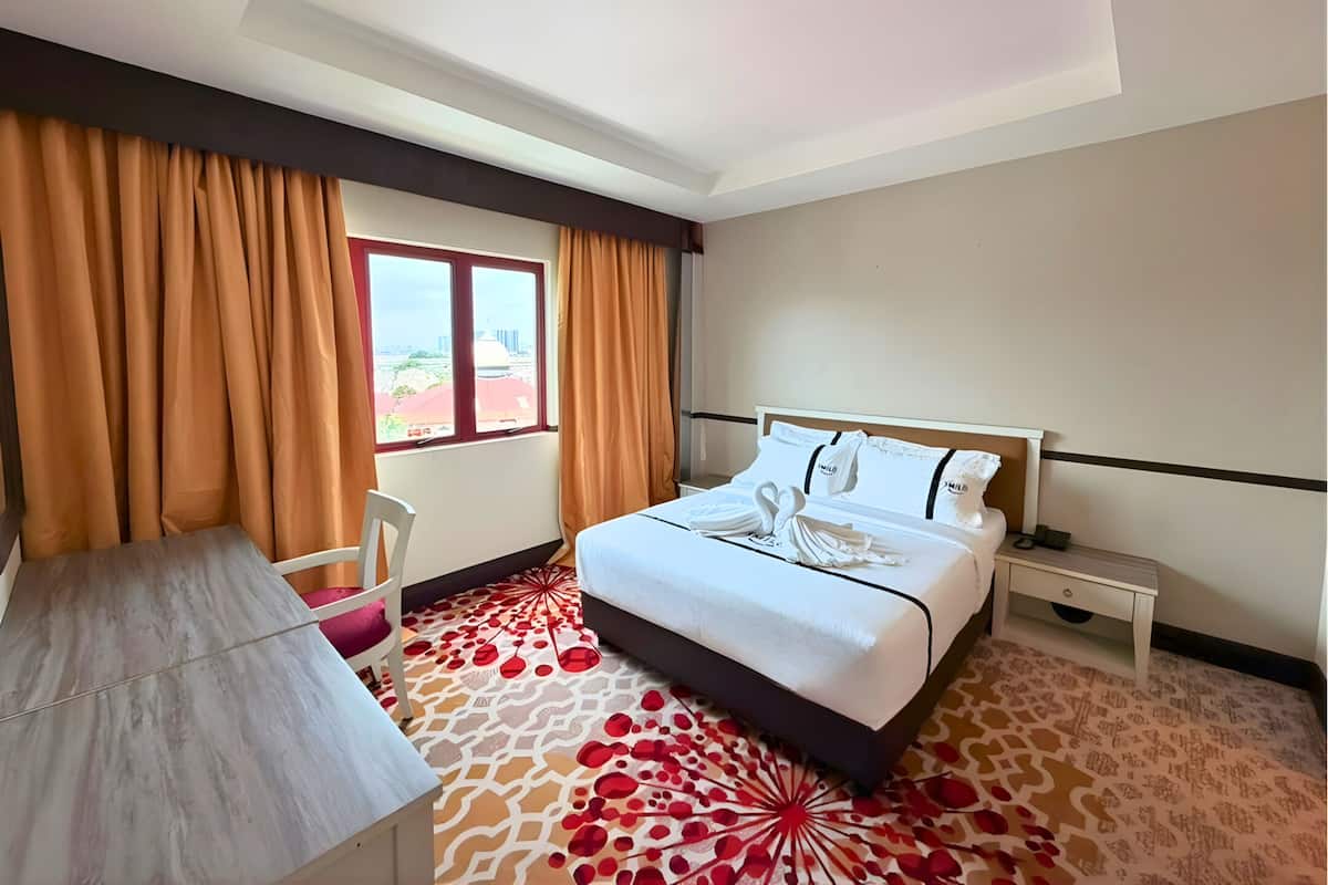 Ein Hotelzimmer mit einem Bett, einem Schreibtisch und einem Sessel. Es gibt ein Fenster mit Vorhängen, und der Boden ist mit einem gemusterten Teppich bedeckt.