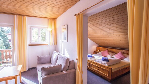 A room with a wooden ceiling, a bed with pillows, a sofa, a table, and a window with curtains. - Chalamandrin Hotel und Restaurant (Scuol)