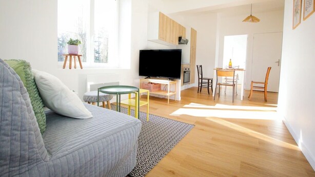A modern living room with a grey sofa, a flat-screen TV, a small table, and a dining area with chairs. - LES LOGIS DU GENERAL - 1st floor apartment (Beaupréau-en-Mauges)