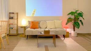 A living room with a sofa, a coffee table, a bookshelf, a lamp, a plant, and a projection screen displaying a building. - Cozy studio with washing machine & dryer in the heart of Hà Nội (Hà Nội)