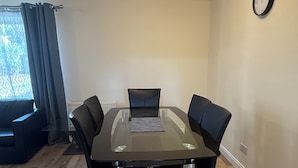 Un coin repas comprenant une table avec un plateau en verre, quatre chaises, une horloge au mur et une fenêtre avec des rideaux.