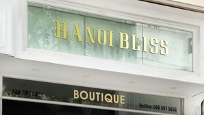 Een etalage met de naam "HANOI BLISS" in gouden letters op een glazen ruit. Daaronder is het woord "BOUTIQUE" te zien, samen met een telefoonnummer "Hotline: 088 697 5656".