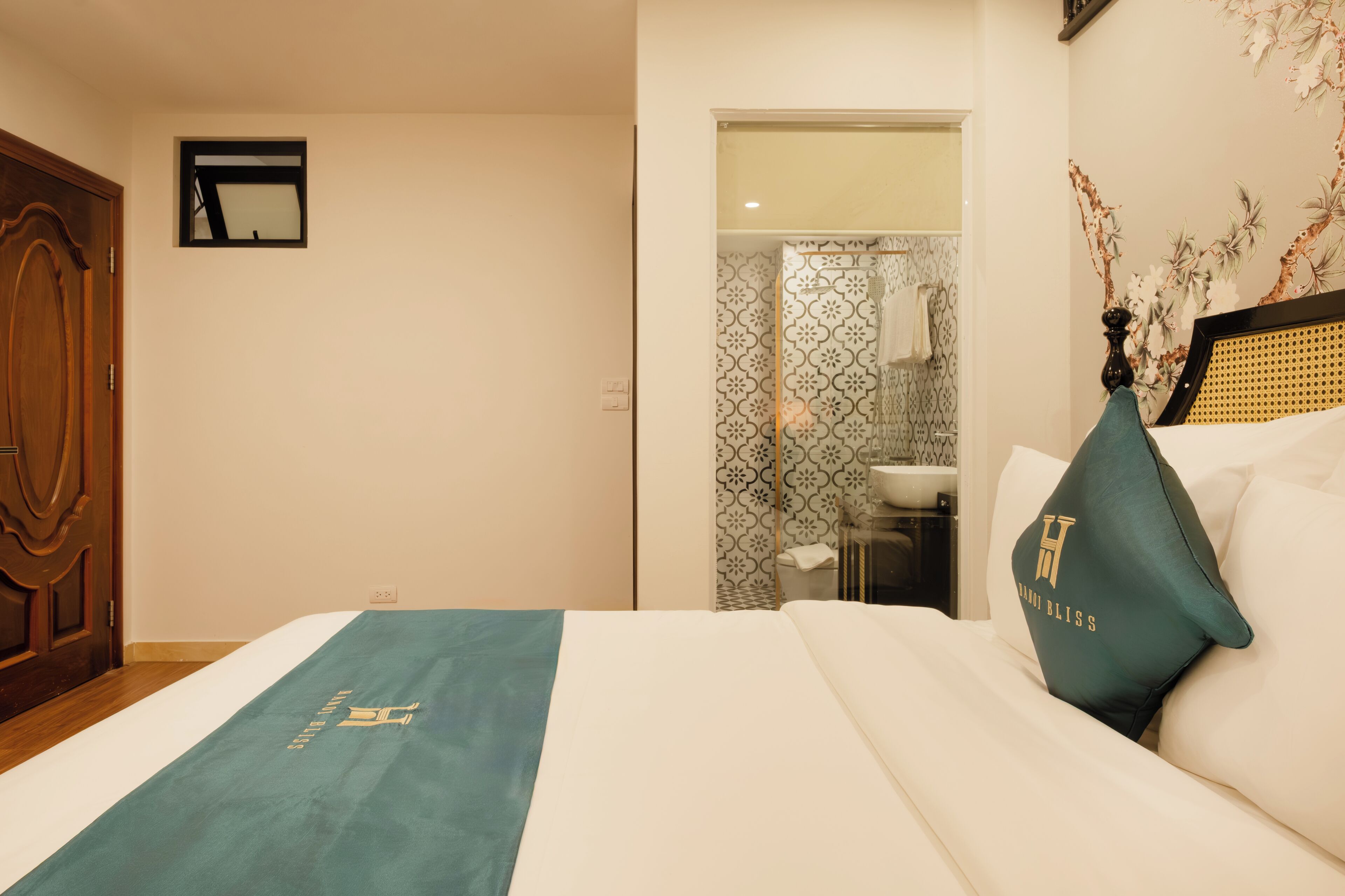 Une chambre d’hôtel avec un lit, une porte en bois, une salle de bain avec douche et un mur orné d’un motif floral.