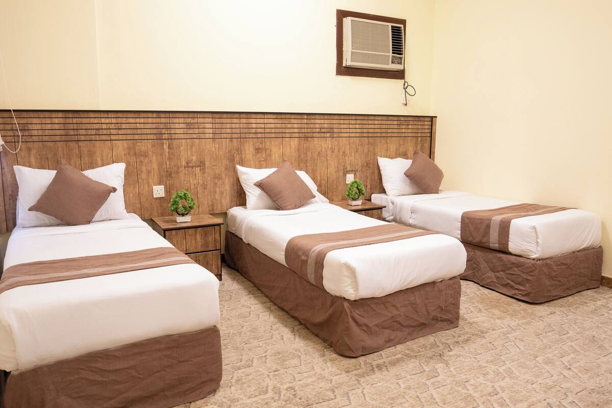 Ein Hotelzimmer mit drei Einzelbetten, jedes mit einer braunen Bettdecke und weißen Kissen. Auf dem Nachttisch neben jedem Bett steht eine kleine Pflanze.
