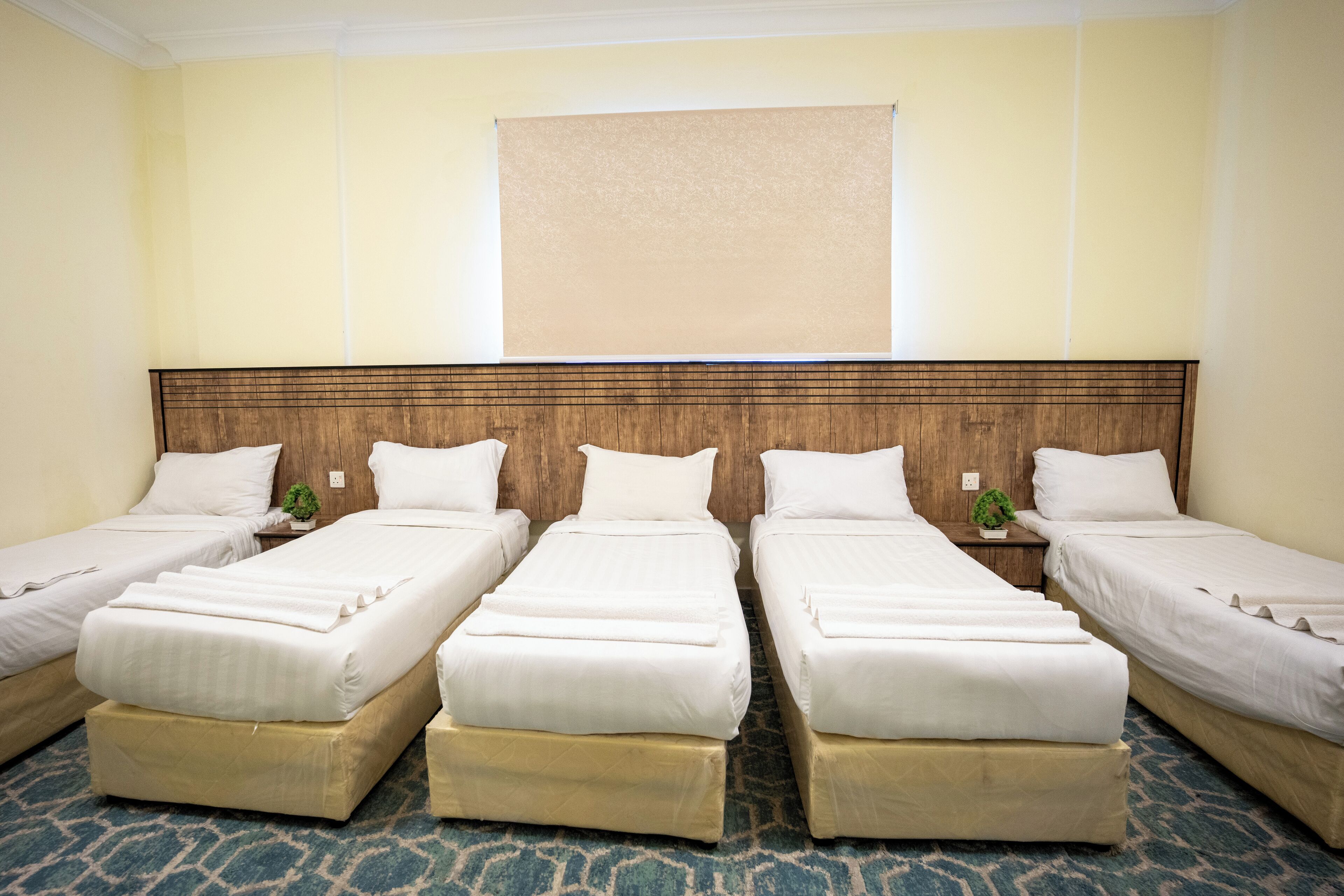 Ein Hotelzimmer mit vier Einzelbetten, einem Holzkopfende, einem großen Fenster mit Jalousien und einem gemusterten Teppich.