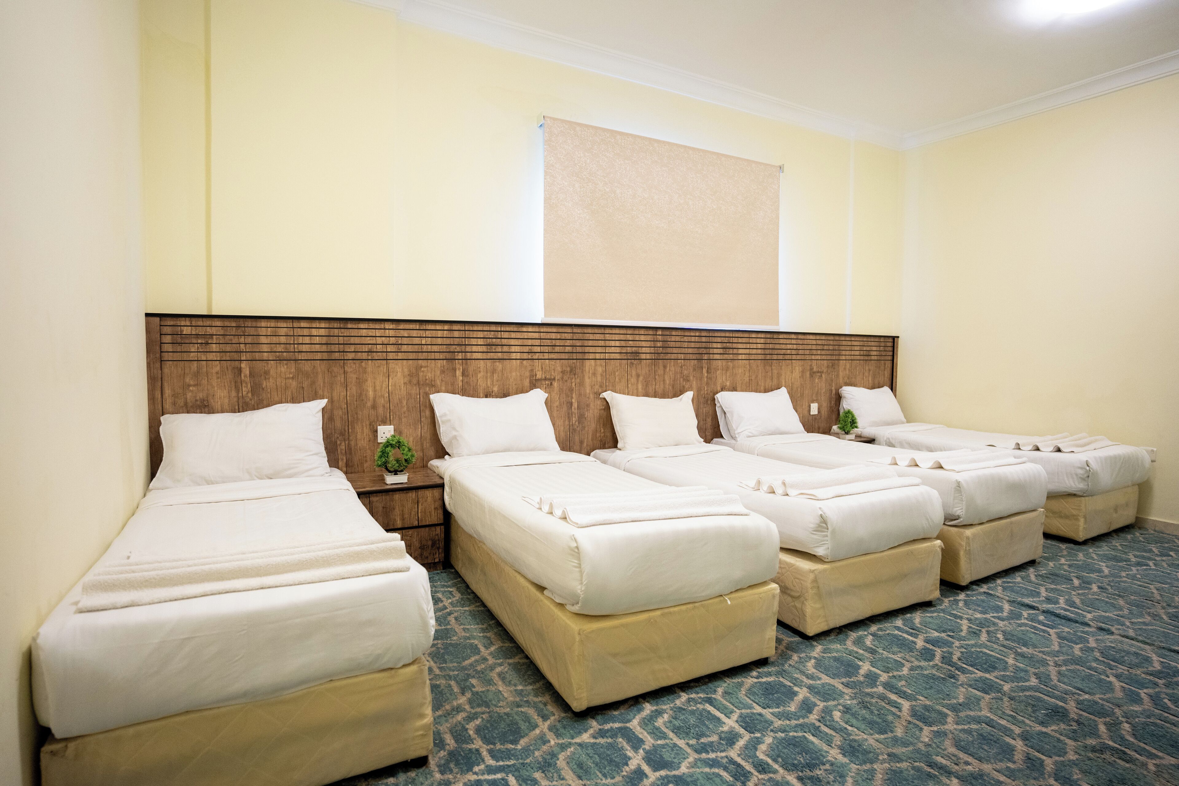 A hotel room with four single beds arranged in a row, a wooden headboard, a beige carpet, and a large window with a beige blind.