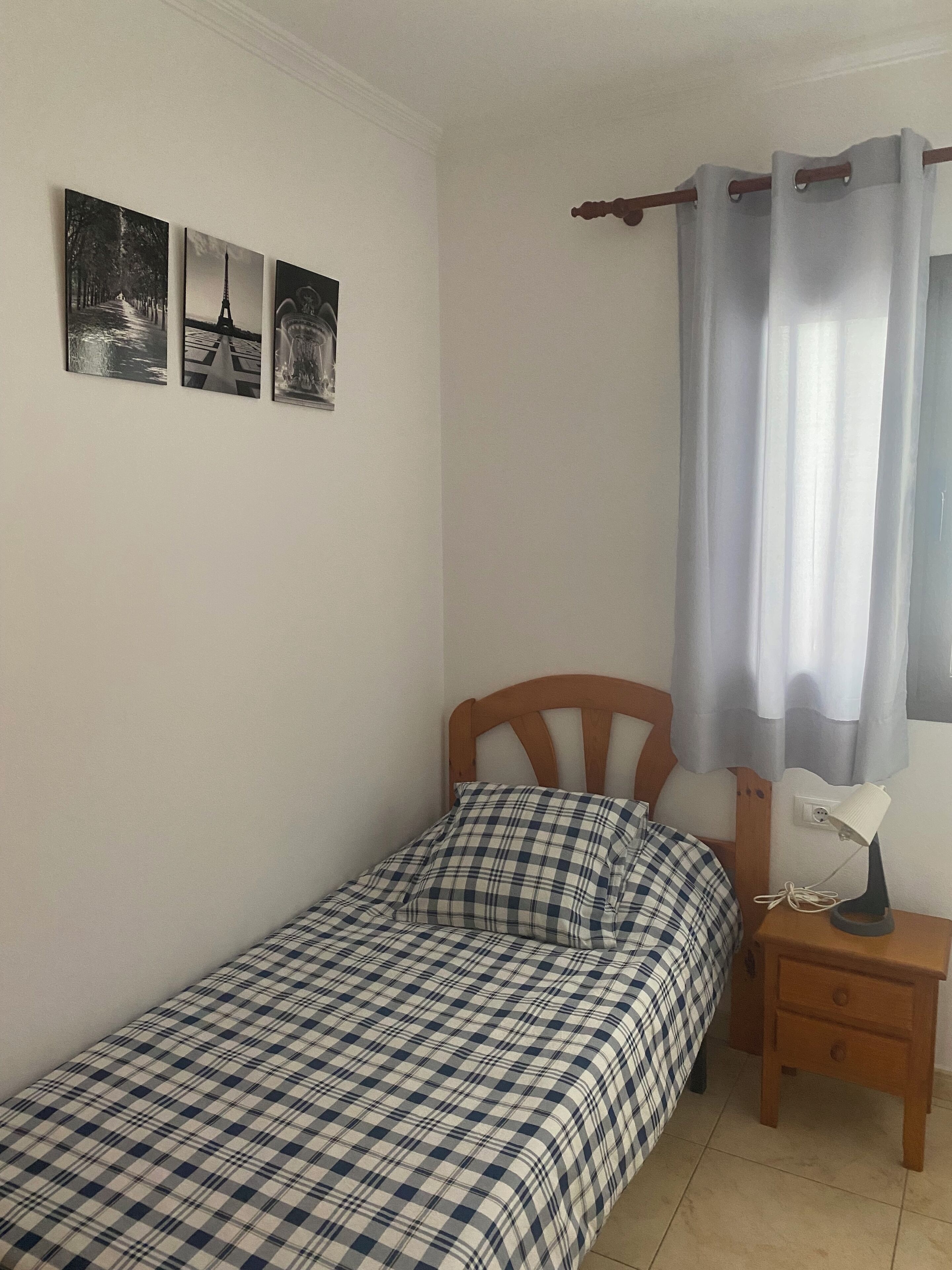 A single bed with a checkered bedspread, a wooden headboard, a nightstand with a lamp, and three framed pictures on the wall.