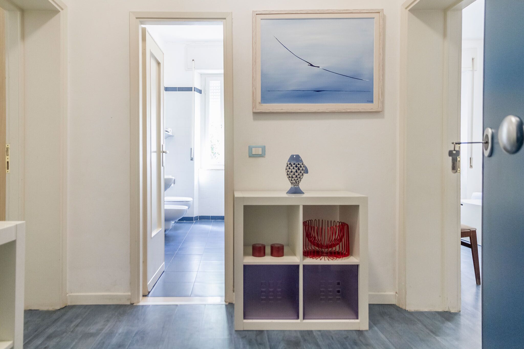 Un corridoio con una porta bianca, un quadro appeso al muro e un piccolo mobiletto bianco con ripiani.