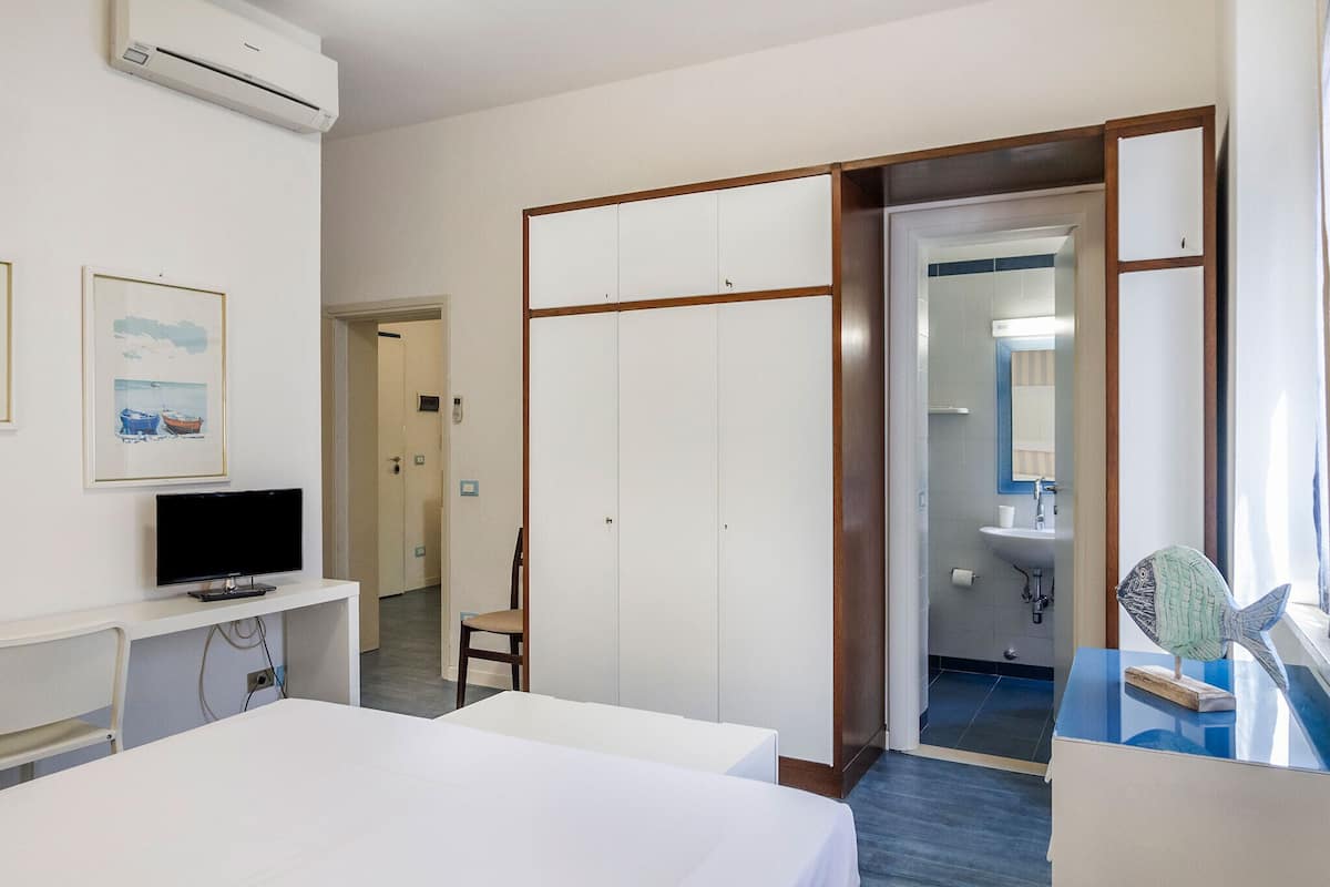 A hotel room with a bed, a desk with a TV, a chair, a wardrobe, and a bathroom visible through an open door.