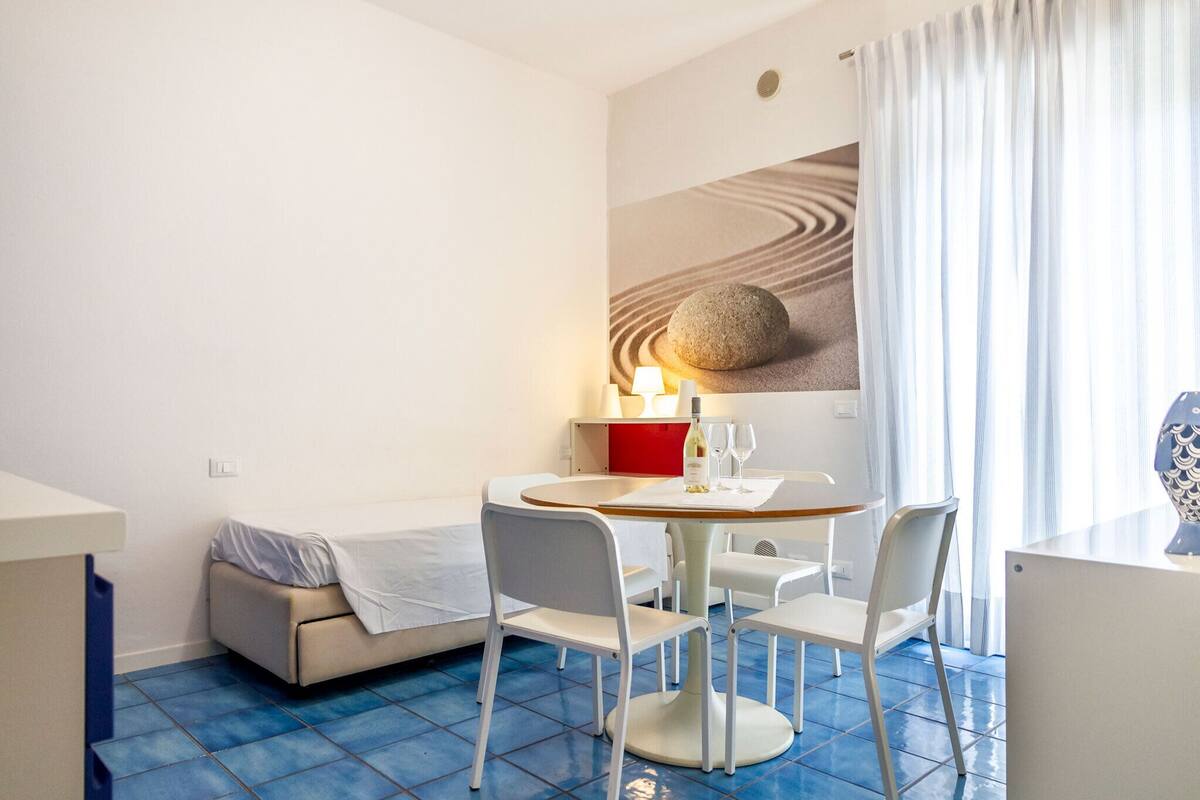 A small, modern room with a round dining table, white chairs, a sofa, and a bed.