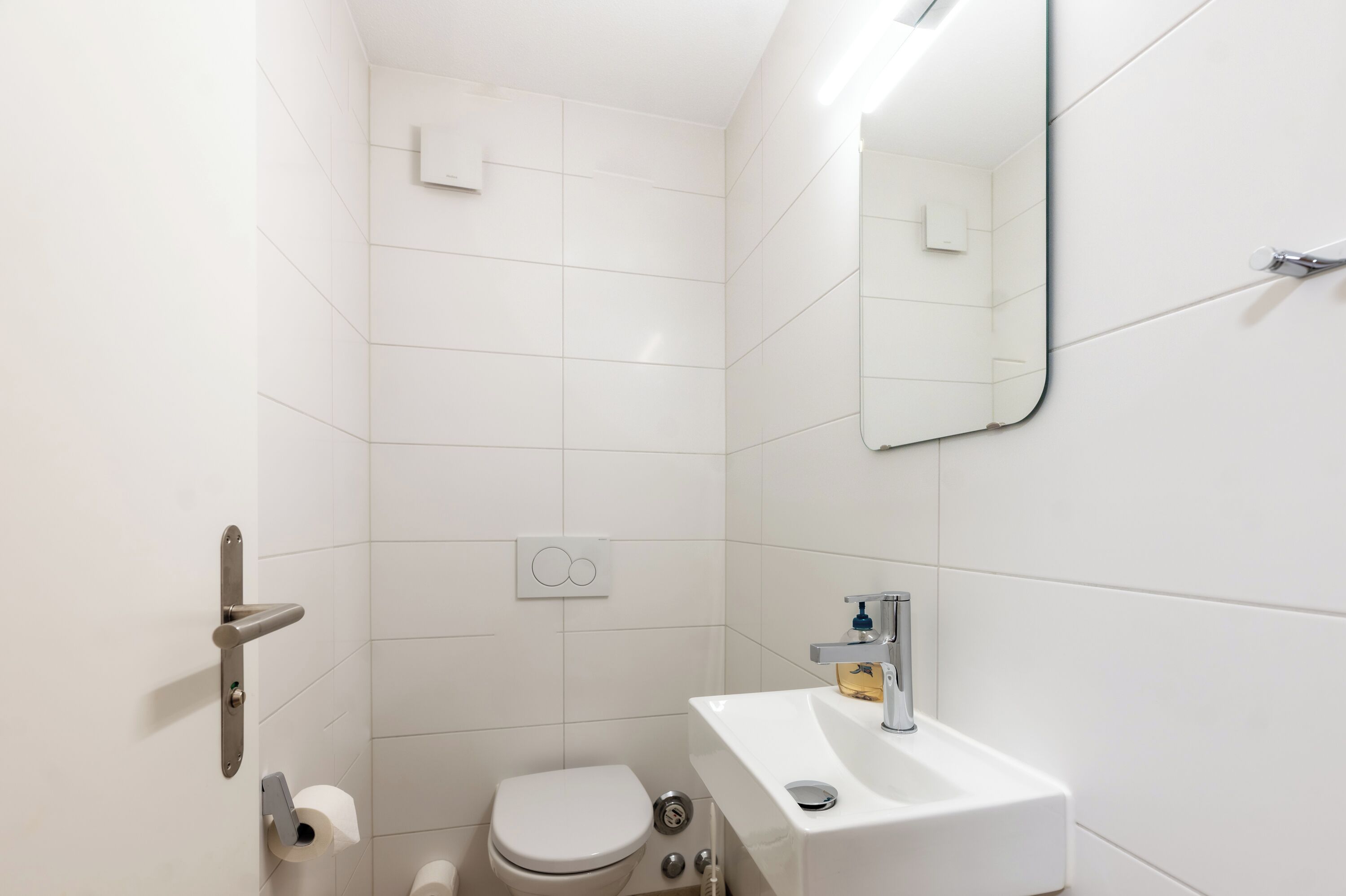 Ein modernes Badezimmer mit weißen Fliesen, Toilette, Waschbecken mit Spiegel und Duschbereich.