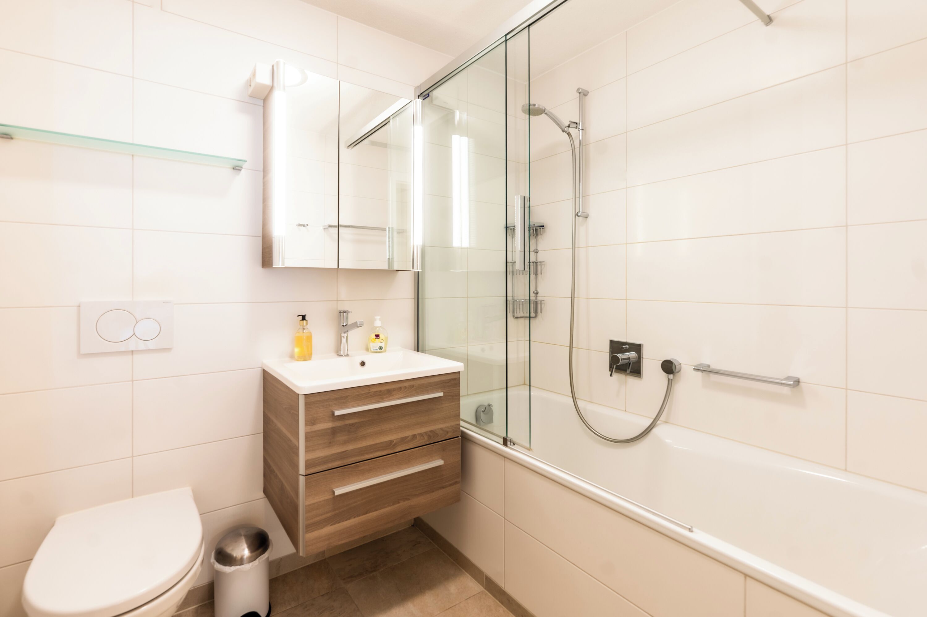 A modern bathroom with a glass-enclosed shower, a white toilet, a wooden vanity with drawers, and a glass shelf above the sink.