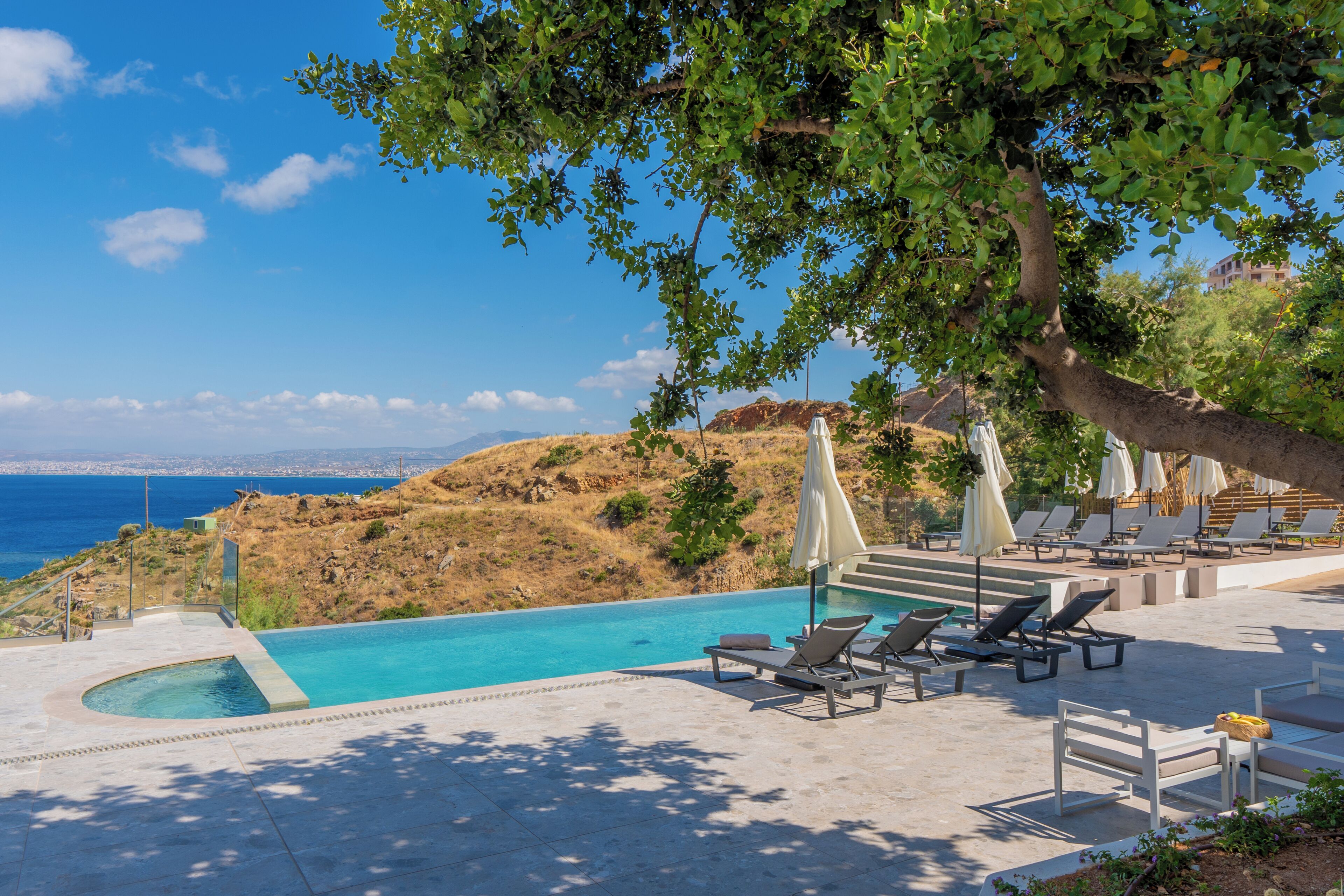 A pool area with lounge chairs and umbrellas, overlooking a body of water and hills.