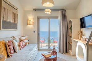 A modern hotel room with a sofa, a table with a book, a TV, and a balcony with red chairs.