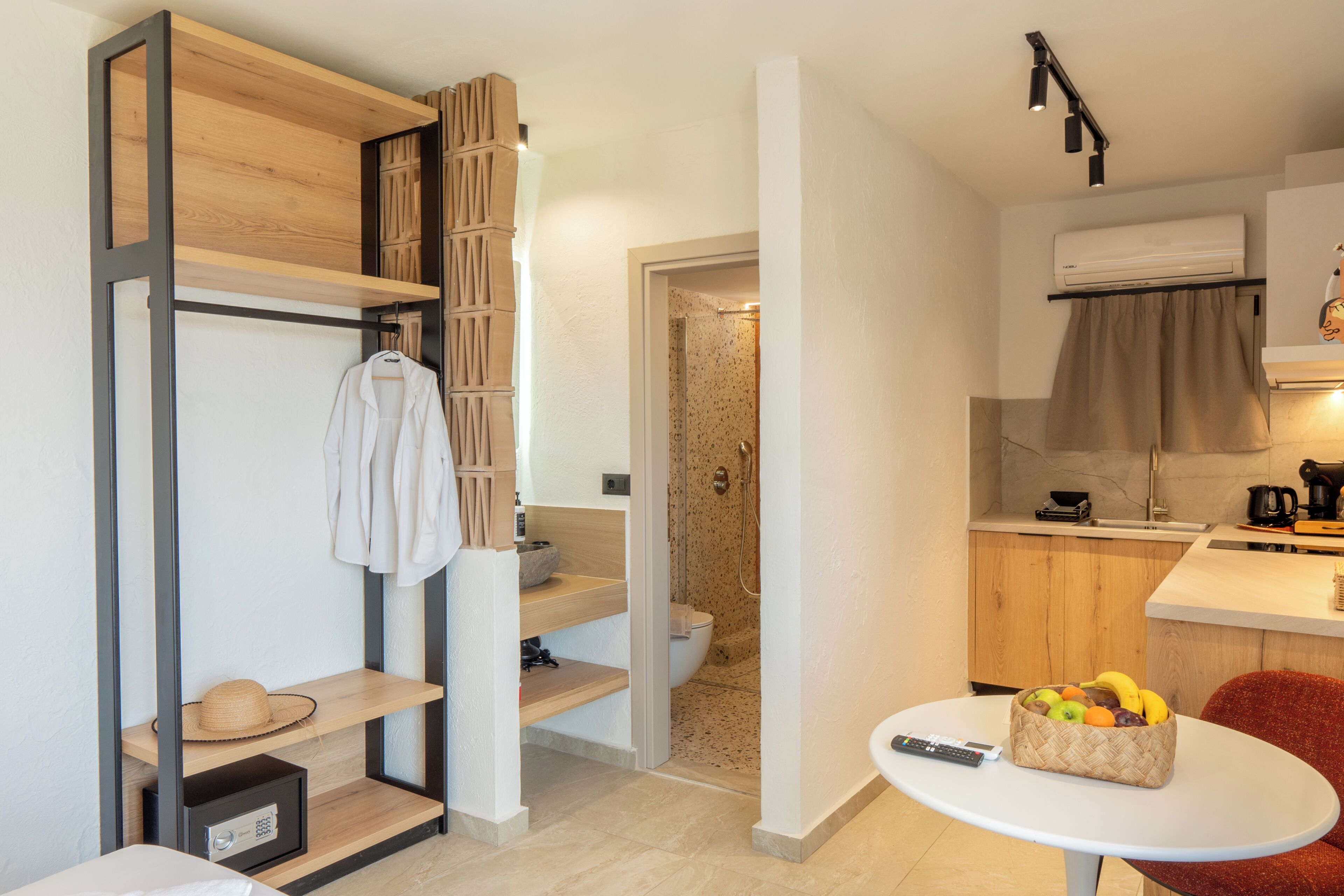 A compact hotel room with a kitchenette, a shower area, and a dining table with fruit.