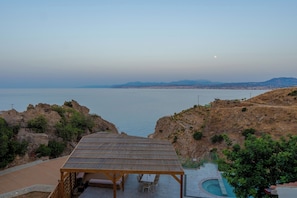 A wooden pergola with a bench and table, overlooking a body of water with a distant cityscape and mountains. - Soluna Forte Sea Living Suites (Malevizi)