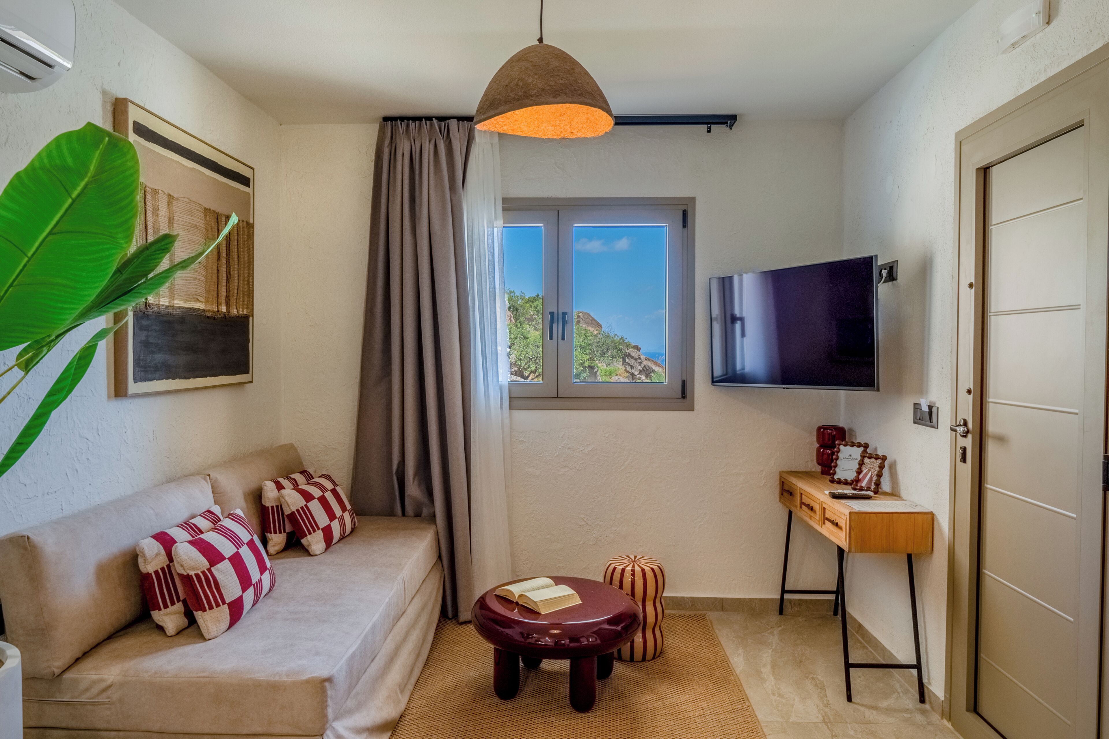 A room with a sofa, a small table, a television, a door, and a window with a view.