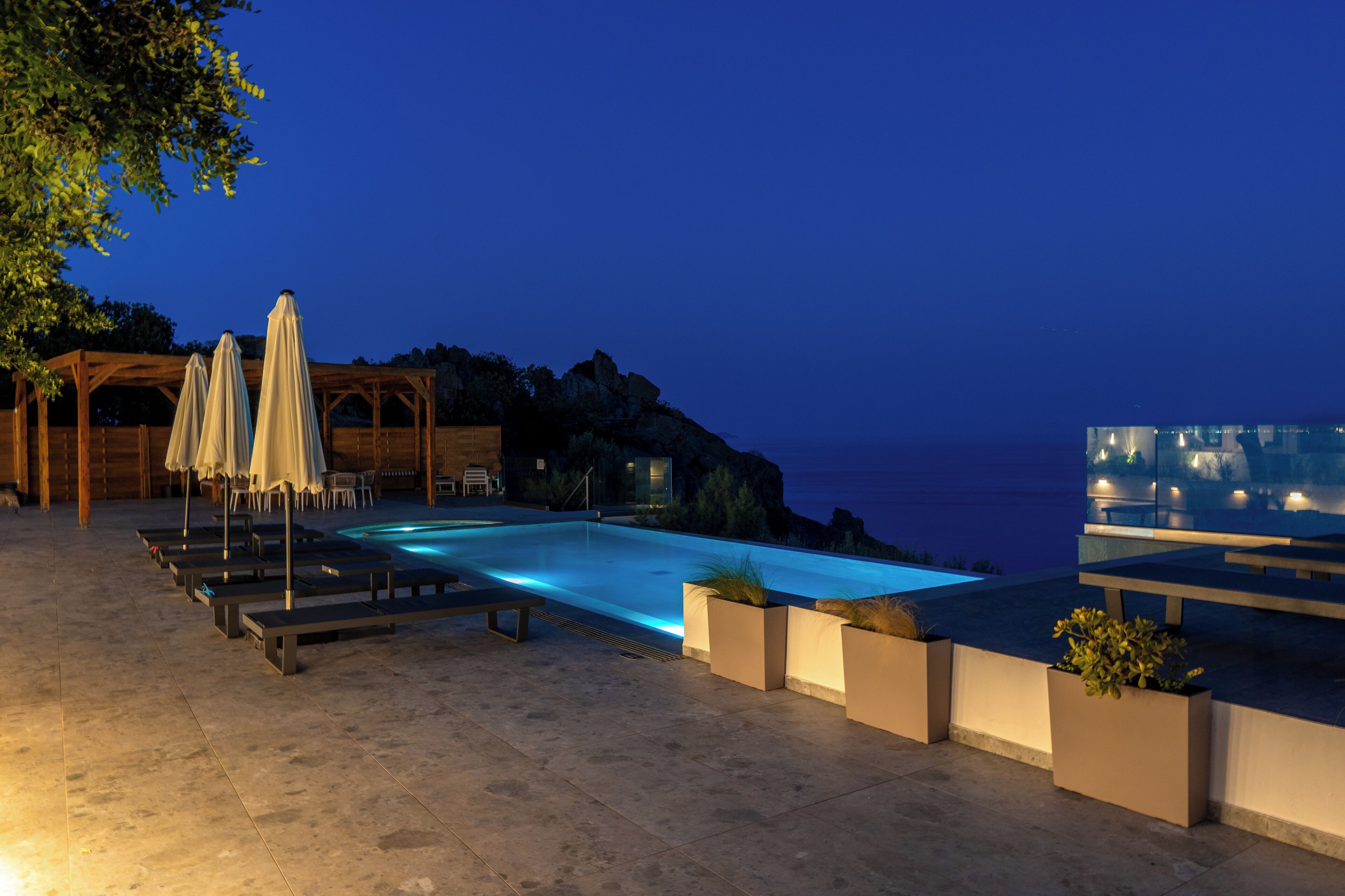 A swimming pool with lounge chairs and umbrellas, a wooden pergola, and a view of the sea at dusk.