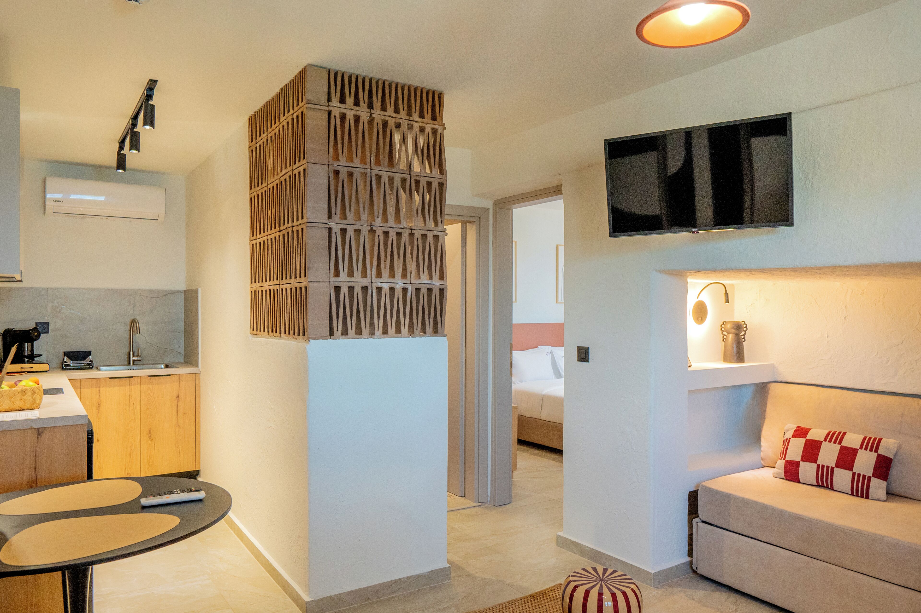 Une chambre d’hôtel moderne dotée d’un coin cuisine, d’un téléviseur fixé au mur, d’un canapé agrémenté d’un coussin à motifs et d’une petite table avec une télécommande.