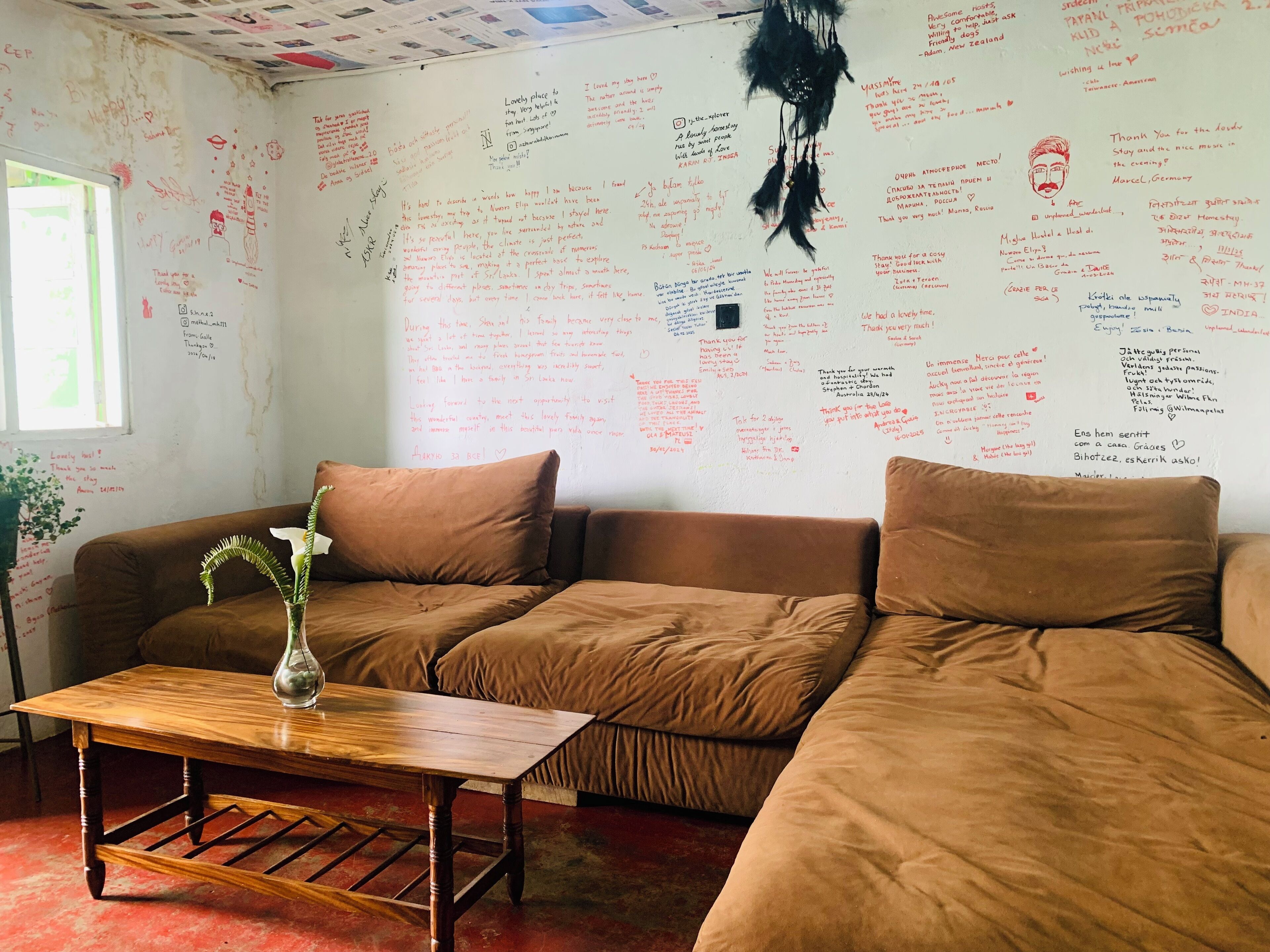 A cozy living room with a brown sofa, a wooden coffee table, and a vase with flowers. The walls are covered with handwritten notes and drawings.