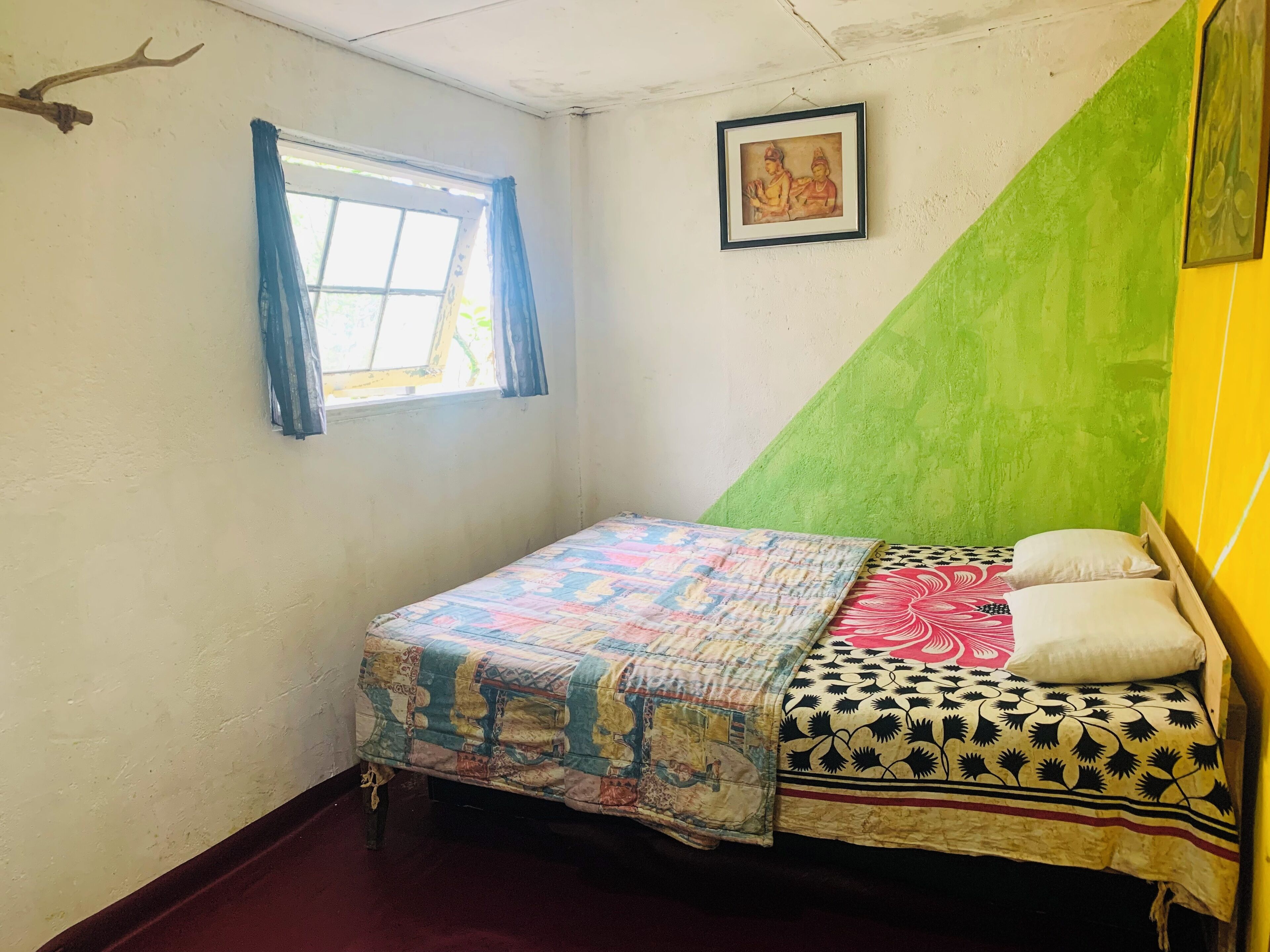 Ein kleines Zimmer mit einem Einzelbett, einer farbenfrohen Bettdecke, einem Fenster mit blauen Vorhängen, einem gerahmten Bild an der Wand und einer Wand in Grün- und Gelbtönen.