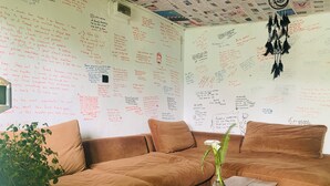 Una sala de estar acogedora con un sofá marrón, una mesa de centro de madera y una planta en maceta. Las paredes están cubiertas de notas y dibujos hechos a mano.
