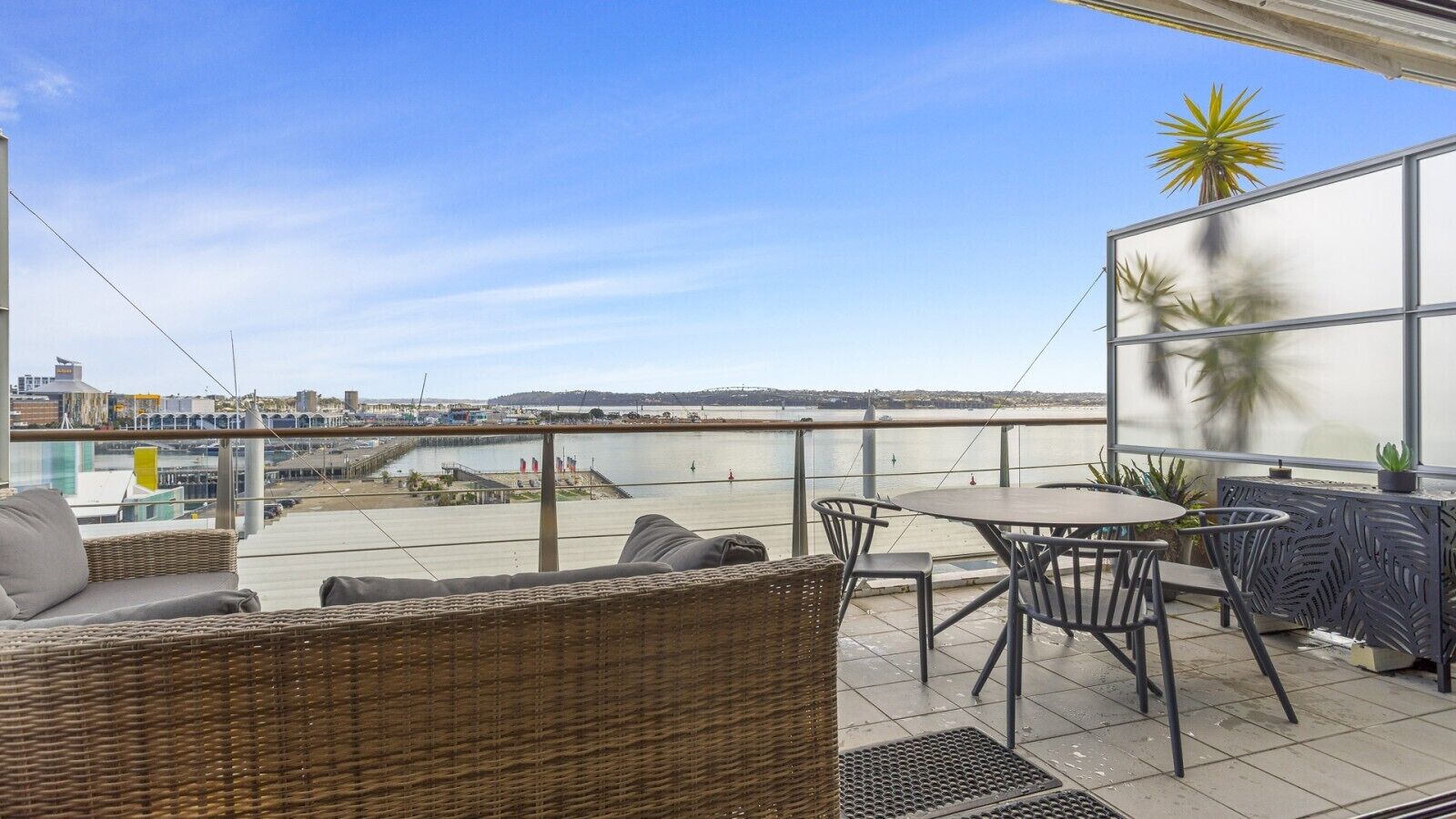A terrace with wicker furniture, a table, and chairs, overlooking a body of water and a distant shoreline.