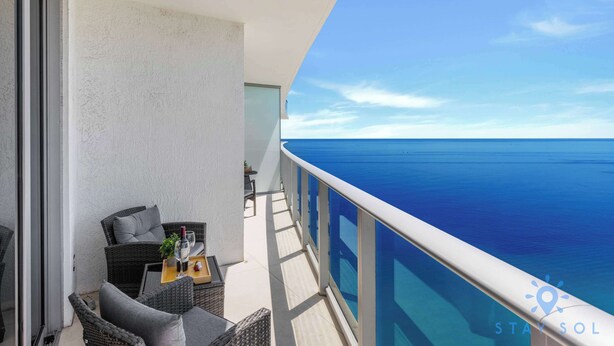 A balcony with wicker furniture and a table, overlooking a clear blue sea. - Hollywood Oceanfront Apt - 2BR (Hollywood)