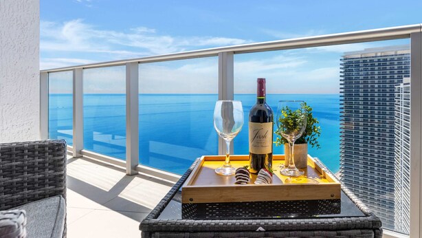 A balcony with a glass balustrade overlooking a clear blue sea, a bottle of wine, and two glasses on a tray. - Hollywood 2BR Apartment w/Balcony (Hollywood)