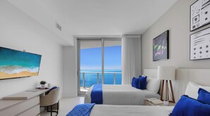 A modern hotel room with two beds, a desk, and a large window offering a sea view.