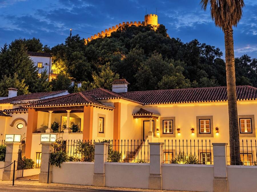 A hotel with a traditional architectural style, featuring a courtyard, a staircase, and a view of a hill with a castle-like structure in the background.